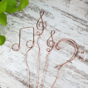 May include: Four copper wire music note shapes on a white wooden background. The shapes include a treble clef, a bass clef, a quarter note, and a half note.