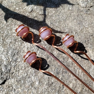 May include: Four copper hair pins with light pink, rounded gemstone beads. The beads are wrapped with copper wire, and the pins have a decorative swirl design. The pins are arranged diagonally on a textured, gray surface.