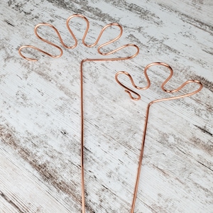 May include: Two copper wire sculptures in the shape of stylized hands. The sculptures are on a white wooden background.