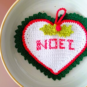 Op de afbeelding: Een wit gehaakt hartvormig ornament met een groene en rode rand. Het ornament heeft het woord "NOEL" in roze garen geschreven. Een groene en gele gehaakte strik is aan de bovenkant van het ornament bevestigd.