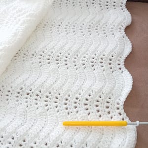 Peut inclure: Une couverture pour bébé en crochet blanc avec un motif ondulé. La couverture est pliée en deux et un crochet à crochet jaune est posé dessus.