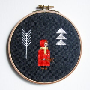 May include: Embroidered hoop art featuring a girl in a red hooded coat and a yellow scarf, standing in a snowy forest with a white tree and a white branch. The background is dark blue.