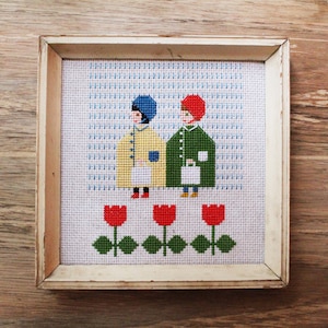 May include: Cross-stitch embroidery framed in a white wood frame. The design features two figures in coats, one in yellow and one in green, standing in front of a row of red tulips.