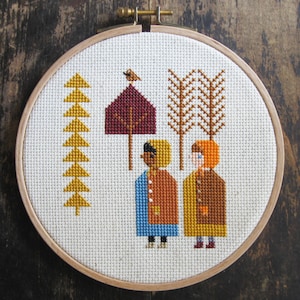 May include: A cross-stitch embroidery hoop depicting a winter scene. Two figures wearing red, orange, and blue coats stand in front of a burgundy house with a bird on top. A row of yellow trees and a brown tree with yellow branches are in the background.