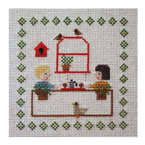May include: Cross-stitch pattern featuring two people sitting at a table with a teapot and cups. There is a birdhouse, a window with a plant, and birds on top of the window. The background is a green and white checkered pattern.