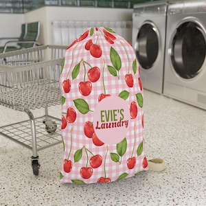 May include: A drawstring laundry bag featuring a pink and white gingham pattern with a repeating cherry and green leaf print. A central pink circle contains the text Evie's Laundry in green and red lettering. The bag has a cinched top closure.