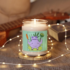 Puede incluir: Una vela de soja en un tarro de vidrio transparente con una etiqueta verde claro que presenta una rana morada de dibujos animados meditando con una zanahoria en una mano y una flor de loto en la otra. La vela está encendida y se encuentra sobre un posavasos de madera.