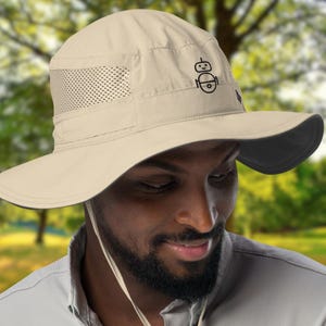 May include: Beige sun hat with a wide brim and a black underside. The hat features a mesh panel for ventilation and a black graphic on the front. It has a chin strap and is designed for sun protection.