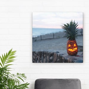 Op de afbeelding: Een uitgesneden ananas met een pompoen-gezicht staat op een stenen muur. De ananas is van binnenuit verlicht en geeft een oranje gloed. De achtergrond toont een strandscène met een hek en oceaan onder een pastelkleurige lucht.