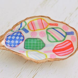 May include: A white seashell with a colorful design of paddleball rackets. The rackets are decorated with stripes, polka dots, and checkered patterns in shades of blue, green, pink, red, and yellow.