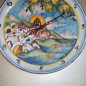 Può includere: Un orologio rotondo in ceramica con una scena costiera dipinta a mano. Il quadrante presenta un cielo blu, edifici bianchi e frutta gialla. Le lancette nere indicano l'ora. L'orologio ha un diametro di circa 25 cm.