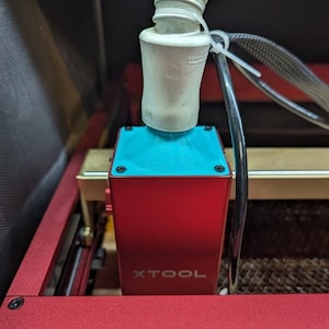 May include: Close-up of a red laser cutter with a white exhaust hose attached to a blue and red box. The box has the word "XTOOL" printed on it. The laser cutter is on a wooden surface.