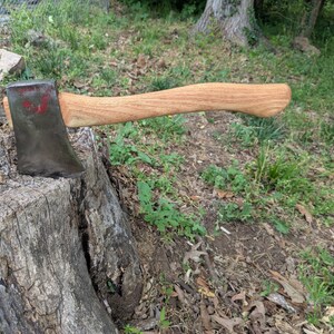 Puede incluir: Hacha con mango de madera y cabeza de metal, apoyada sobre un tocón de árbol. La cabeza del hacha tiene un aspecto oscuro y desgastado con marcas rojas. El mango de madera es de color marrón claro y curvado, mostrando la veta de la madera. El hacha está colocada horizontalmente.