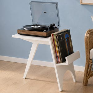 Op de afbeelding: Een witte houten bijzettafel met schuine poten en een geïntegreerd opbergvak voor vinylplaten. Een donkerhouten platenspeler met een doorzichtige kunststof stofkap staat op het tafelblad, terwijl een verzameling vinylplaten rechtop is geplaatst.
