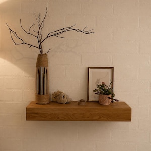 Op de afbeelding: Een rechthoekige zwevende houten plank gemonteerd aan een witte bakstenen muur. Op de plank staat een hoge cilindrische vaas omwikkeld met jutekoord met kale takken, een kleine steen, een ingelijste lijntekening en een kleine groene vetplant.