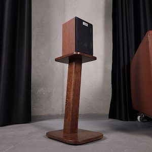 May include: A dark brown wooden speaker stand with a rectangular base and a vertical support column, topped with a square platform. A black and wood-toned speaker with a small white logo on the front grille is positioned on the top platform.