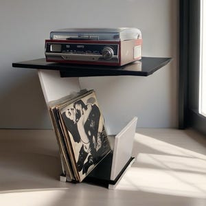 May include: A retro-style record player and vinyl storage unit. The unit has a black and white geometric design with a red and silver record player on top. Vinyl records are stored in a compartment below. The unit is placed near a window.