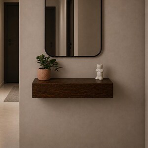May include: A rectangular wall-mounted floating shelf made of dark brown wood with a smooth finish. The shelf is positioned below a rectangular mirror with a thin black frame. A small green plant in a textured pot and a white figurine sit on the shelf.