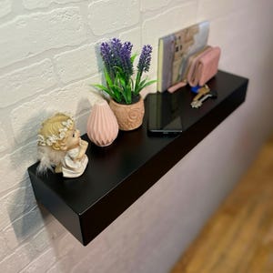 May include: A black floating shelf on a white brick wall, with a small angel figurine, a pink vase, a potted lavender plant, a phone, a wallet, keys, and a small book.