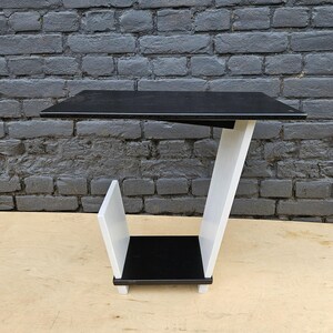 May include: A C-shaped side table featuring a flat black rectangular top surface supported by a slanted white structural panel. The base consists of a small black square shelf positioned above a white vertical divider, creating a storage section.