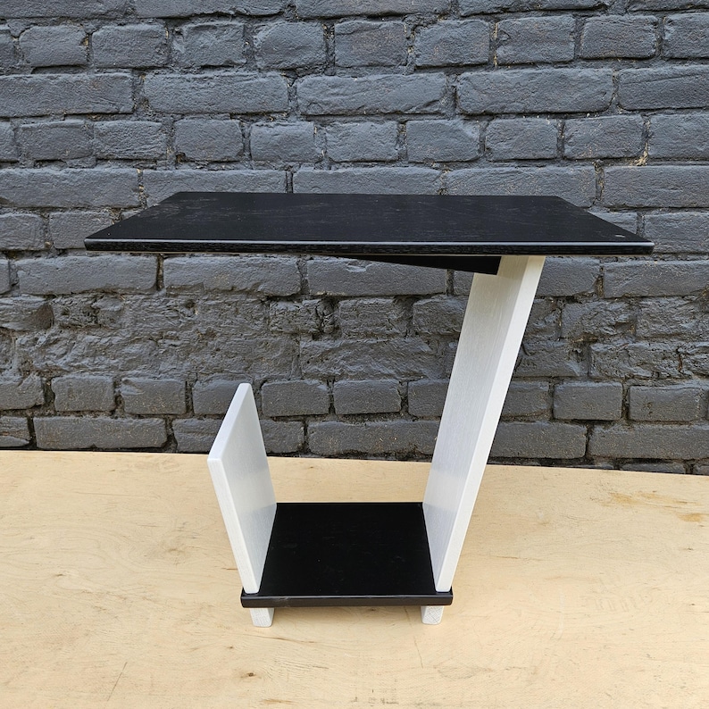 May include: A C-shaped side table featuring a flat black rectangular top surface supported by a slanted white structural panel. The base consists of a small black square shelf positioned above a white vertical divider, creating a storage section.