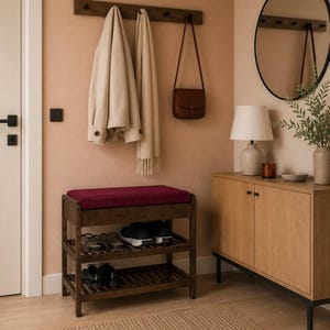 May include: A dark wood shoe rack with a deep red cushioned seat on top and two slatted shelves below. The rack holds several pairs of shoes, including black sneakers and dress shoes, providing a functional storage solution for an entryway.