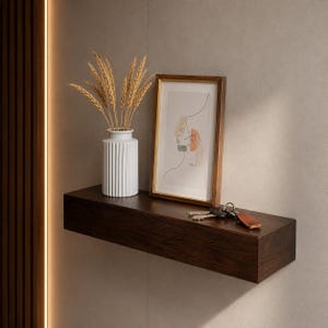 May include: A dark brown wooden floating shelf holding a white fluted vase filled with dried wheat stalks, a framed line art print with a gold border, and a set of metal keys with a brown leather keychain resting on the flat surface.