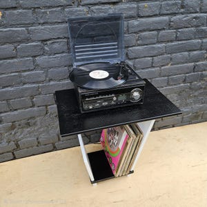 May include: A black record player with a clear plastic lid sits on a black and white side table. The table features a lower shelf holding a collection of vinyl records. The record player has a front control panel with a dial and digital display.