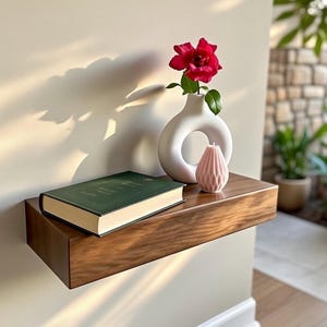 May include: A floating wooden shelf with a book, a white vase with a red flower, and a pink candle. The shelf is mounted on a light-colored wall. The vase has a circular opening. The candle has a geometric design.