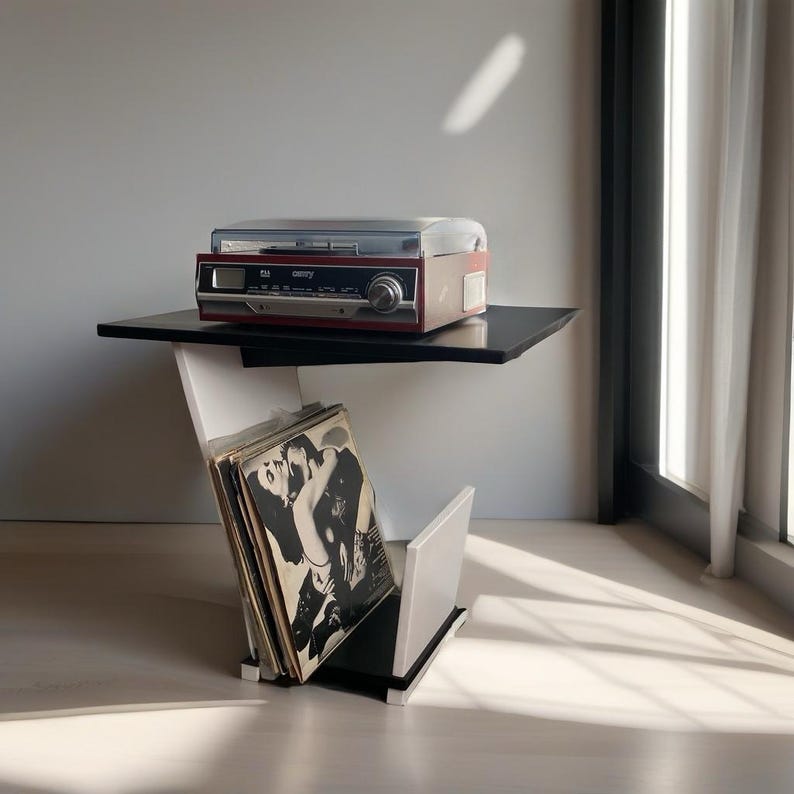 May include: A red and black record player with a clear plastic lid sits on a black tabletop. Below the tabletop, a white and black storage unit holds a collection of vinyl records with black and white illustrated covers in a vertical rack.