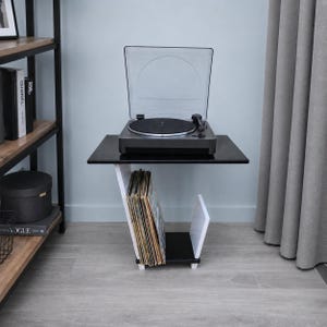 May include: A black and white side table featuring a top surface holding a black turntable with a clear dust cover. The base of the table includes a slanted white support and a black bottom shelf designed to store a collection of vinyl records.
