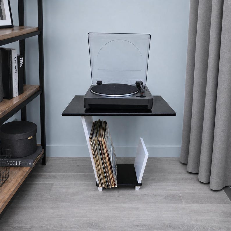 May include: A black and white side table featuring a top surface holding a black turntable with a clear dust cover. The base of the table includes a slanted white support and a black bottom shelf designed to store a collection of vinyl records.