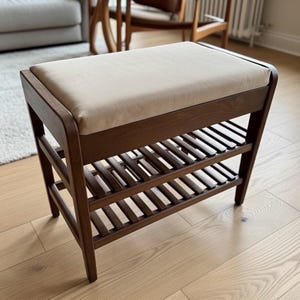 May include: A dark brown wooden shoe bench with a cream-coloured cushioned seat. It features two slatted shelves for storage. The bench is a stylish and functional piece of furniture, suitable for a hallway or bedroom.