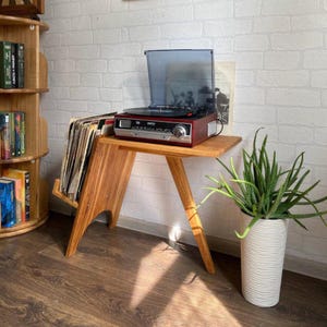 May include: A wooden record player stand with a built-in record holder. A red and silver record player sits on top, with a stack of vinyl records in the holder. A potted plant is to the right, and a wooden bookshelf is to the left.