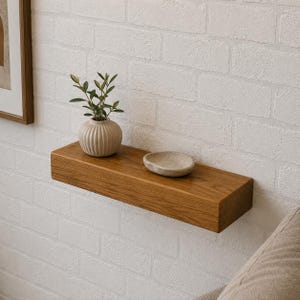 May include: A rectangular floating wooden shelf mounted on a white brick wall. On the shelf sits a beige ribbed ceramic vase containing a small green plant and a shallow, smooth, light-colored stone bowl placed next to the vase.