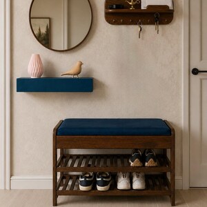 May include: A dark wood shoe bench with a navy blue upholstered seat and two slatted shelves holding four pairs of sneakers. Above it, a blue floating shelf displays a pink geometric vase and a wooden bird figurine near a round mirror.