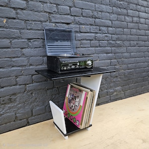 May include: A black and white record player stand featuring a top shelf holding a black turntable with a clear plastic lid and a lower storage section containing a collection of vinyl records with a visible album cover titled Public Enemy.
