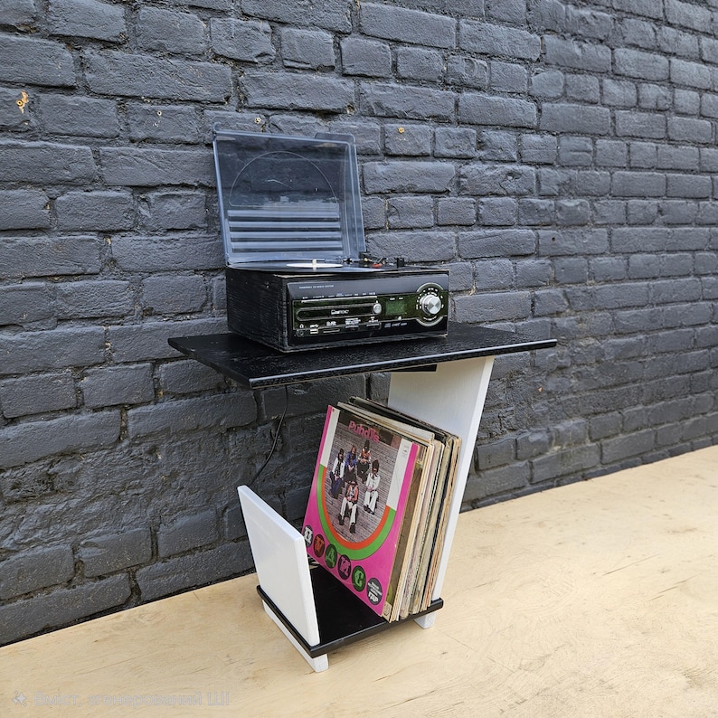 May include: A black and white record player stand featuring a top shelf holding a black turntable with a clear plastic lid and a lower storage section containing a collection of vinyl records with a visible album cover titled Public Enemy.