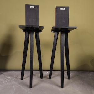 May include: Two black wooden speaker stands, each featuring a tripod base with three angled legs and a flat square top platform. A rectangular black speaker with a silver logo plate is positioned on top of each stand, creating a matching pair.