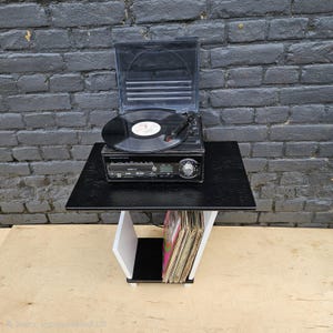 May include: A black turntable with a clear plastic lid and a vinyl record on the platter, resting on a black and white side table. The table features a lower shelf containing a collection of vinyl records stored vertically in a row.