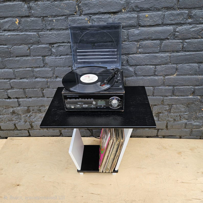 May include: A black turntable with a clear plastic lid and a vinyl record on the platter, resting on a black and white side table. The table features a lower shelf containing a collection of vinyl records stored vertically in a row.