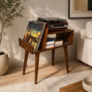 May include: A dark brown wooden record player stand with four tapered legs. The top surface holds a dark wood-toned record player, while the angled side compartment stores vinyl records and the lower shelf holds several stacked hardcover books.