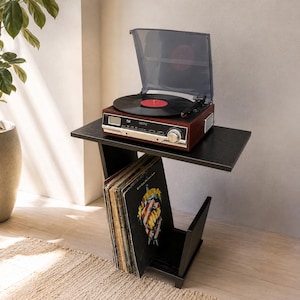 May include: A dark wood record player with a clear plastic dust cover and a black vinyl record sits on a black side table. The table features a lower storage compartment filled with a collection of vinyl records in their sleeves.