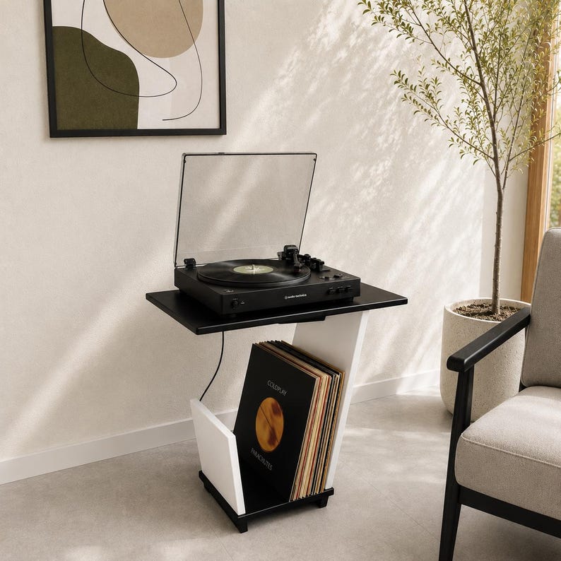 May include: A black turntable with a clear dust cover sits on a two-tone black and white side table. The table features a top surface for the record player and a lower storage compartment containing a collection of vinyl records in sleeves.
