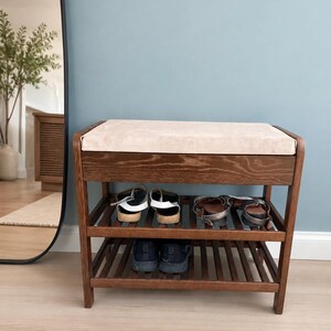 May include: A dark wood shoe bench featuring a rectangular beige upholstered seat cushion on top. The unit has two slatted shelves underneath for storage, currently holding two pairs of sandals on the top shelf and one pair of black sneakers below.