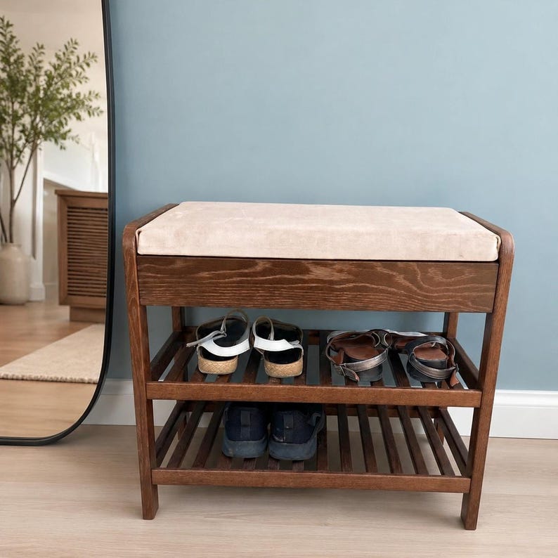 May include: A dark wood shoe bench featuring a rectangular beige upholstered seat cushion on top. The unit has two slatted shelves underneath for storage, currently holding two pairs of sandals on the top shelf and one pair of black sneakers below.