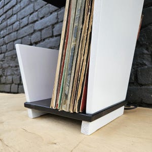 May include: A wooden vinyl record storage rack featuring a black base and white side panels. The rack holds a collection of vintage vinyl records with visible paper sleeves in various colors, standing upright between the two white side supports.