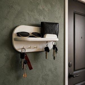 May include: A white wooden key holder with a shelf, three hooks, and a small compartment. The shelf holds sunglasses and a black wallet. Several sets of keys hang from the hooks. The holder is mounted on a green wall.