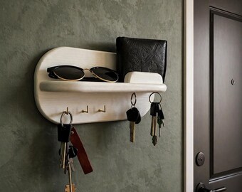 May include: A white wooden wall-mounted key holder with a shelf, three hooks, and a small compartment. The shelf holds a pair of sunglasses and a black wallet. Several sets of keys hang from the hooks. The holder is mounted on a green wall.