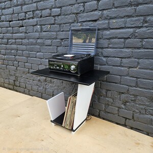 May include: A black and white record player stand featuring a flat top surface for a turntable and a lower angled storage compartment for vinyl records. The structure is constructed with a black top and base with a white angled support panel.